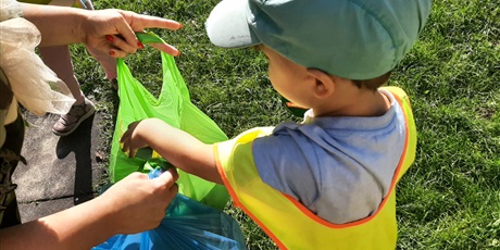 Powiększ grafikę: Chłopiec w kamizelce odblaskowej wrzuca śmieci do zielonego worka na tle trawy w ogrodzie