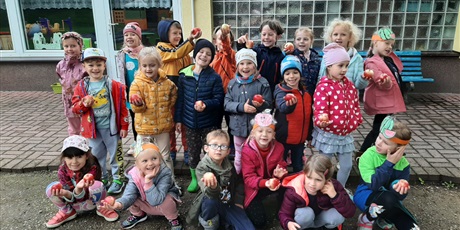 Powiększ grafikę: Grupa 6-latków stoi w ogrodzie przedszkolnym przy tarasie i pokazuje zebrane jabłka